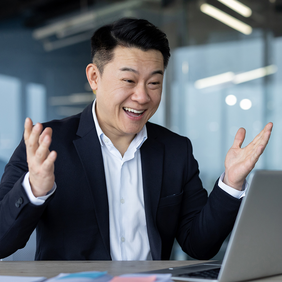 Businessman in a dark suit gesturing with open hands while sitting at a desk with a laptop in a modern office.