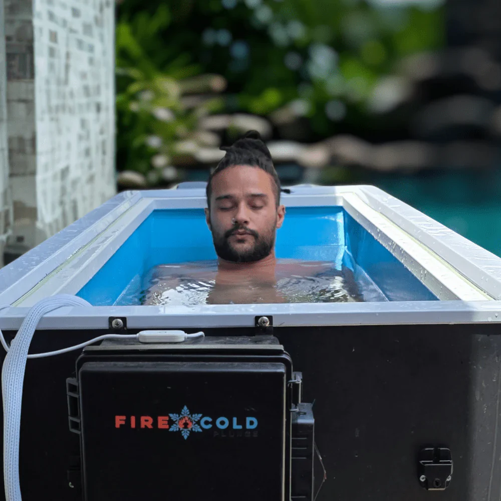 A man with his eyes closed and hair in a bun is submerged up to his chest in a cold plunge tub outdoors. The tub is black with a blue interior and white trim. A control box with the "FIRE & COLD PLUNGE" logo is attached to the front. In the background, lush green foliage and a swimming pool are visible.
