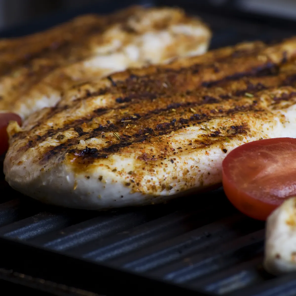 Close-up of seasoned chicken breasts being grilled on a ridged grill pan, with slices of tomato visible on the side.