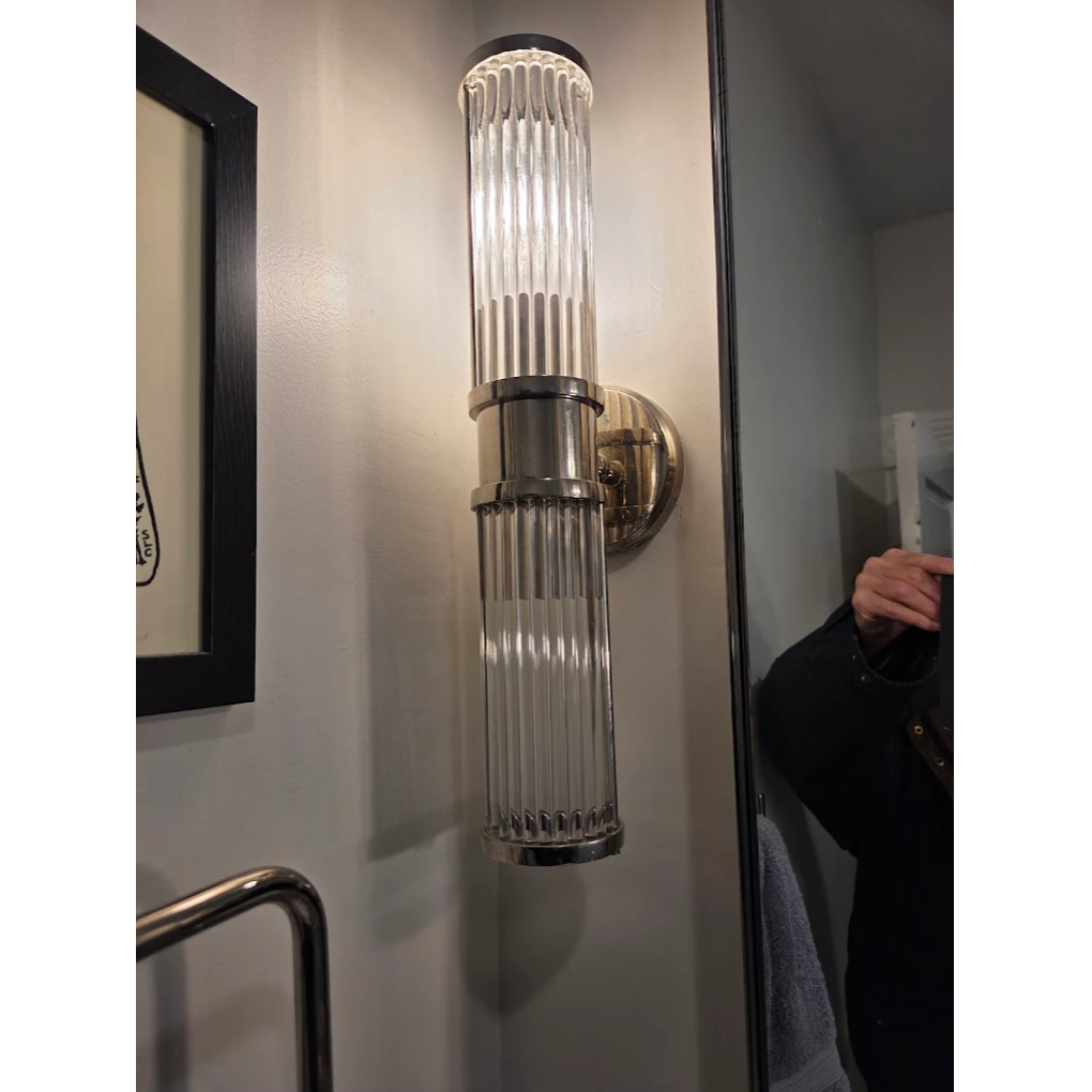 A modern wall-mounted bathroom light fixture with ribbed glass cylinders and a chrome base, illuminated beside a mirror.