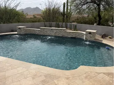 Scottsdale pool remodel with a custom stacked stone spillway wall and dual scupper water features, travertine decking, and desert landscaping as part of a full backyard renovation. Camelback Mountain visible from the pool deck. The stacked stone wall ties the water feature into the overall design rather than looking like an add-on. Built by Hacienda Outdoors, ROC #325952.