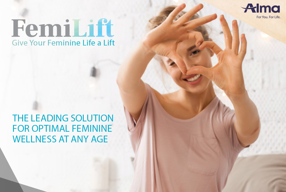 A young woman smiles and forms a heart shape with her hands over her eyes in a bright, airy room with decorative string lights in the background. Text overlays announce "FemiLift: Give Your Feminine Life a Lift" and "THE LEADING SOLUTION FOR OPTIMAL FEMININE WELLNESS AT ANY AGE." The Alma logo is in the upper right corner.