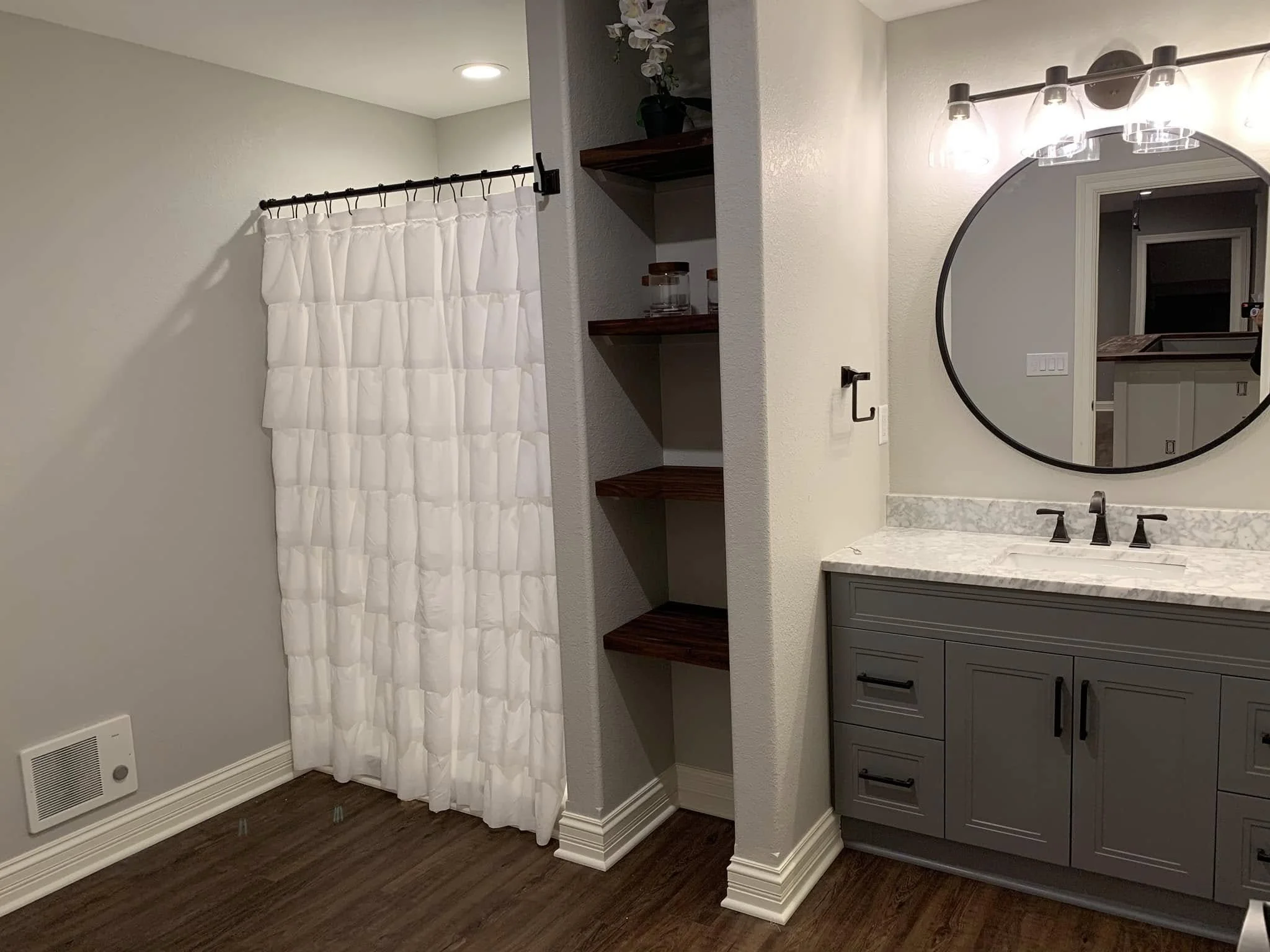 A modern bathroom features a gray vanity with a white marble countertop and a round mirror above it. A white ruffled shower curtain hangs on the left, and a built-in open shelving unit with dark wood shelves is in the center. The floor is dark wood.