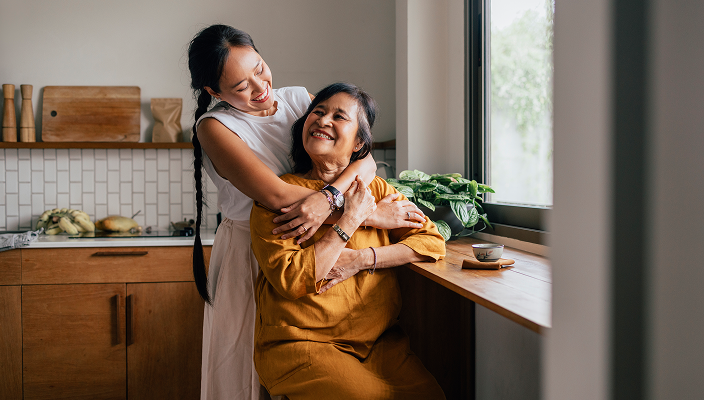 A mother and her adult daughter hugging in the kitchen.