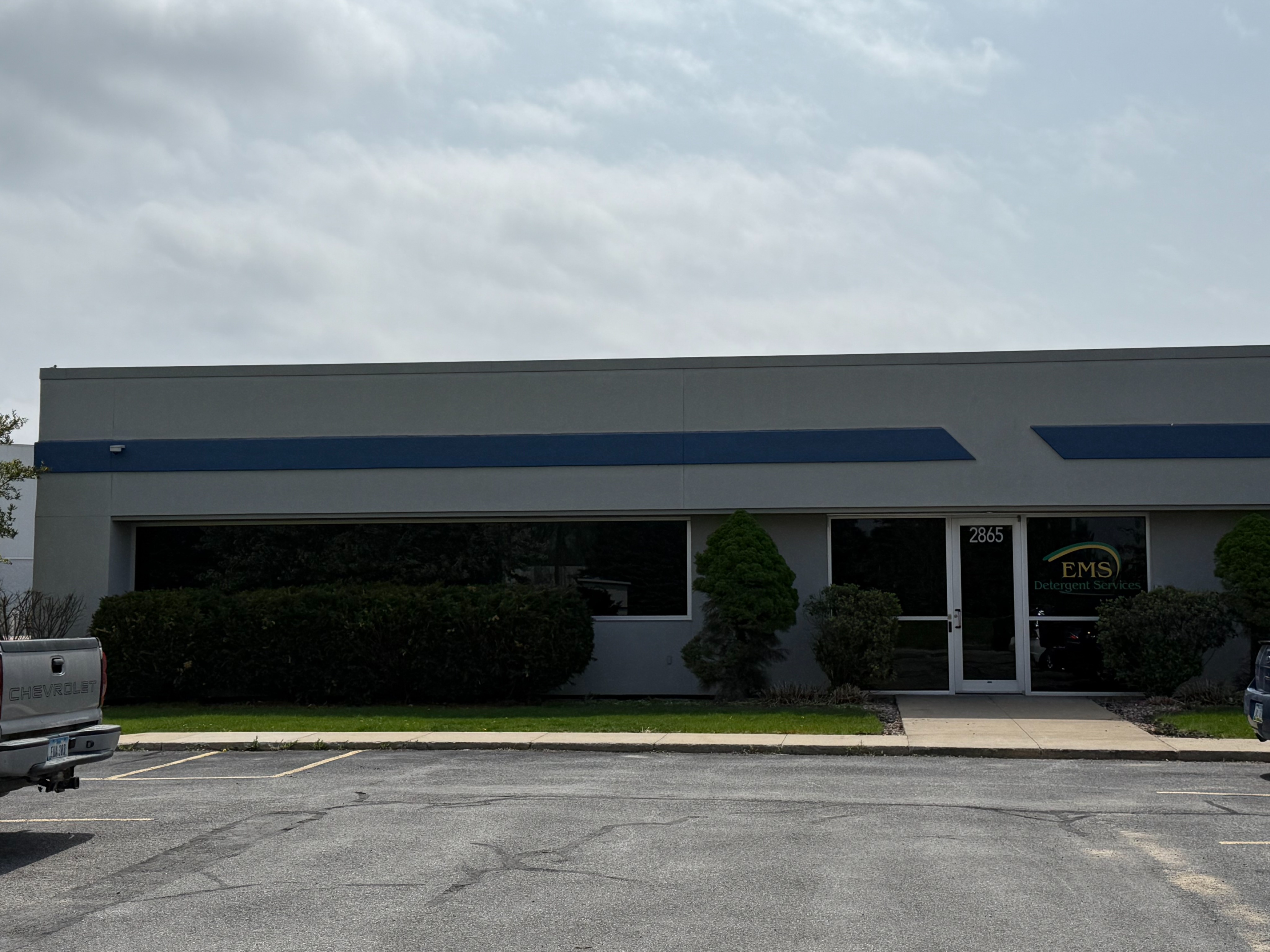 A single-story commercial building with a clean, modern exterior houses EMS Detergent Services, with parking spaces and trimmed landscaping in front under a lightly overcast sky.