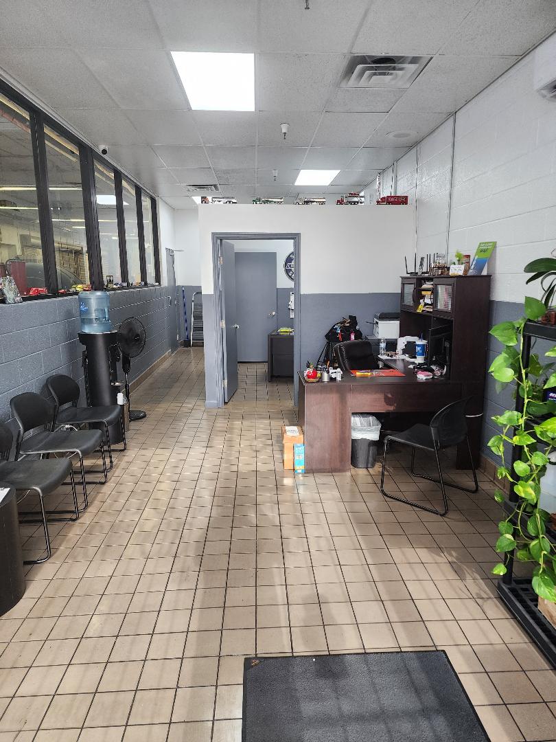 A well-lit, tiled waiting area with black chairs arranged along a brick wall. A water cooler with a blue bottle is next to a fan. In the background, a wooden desk is set up with a computer and office supplies. Several small toy trucks are displayed on a shelf above the desk. Plants in pots add a touch of greenery to the scene.