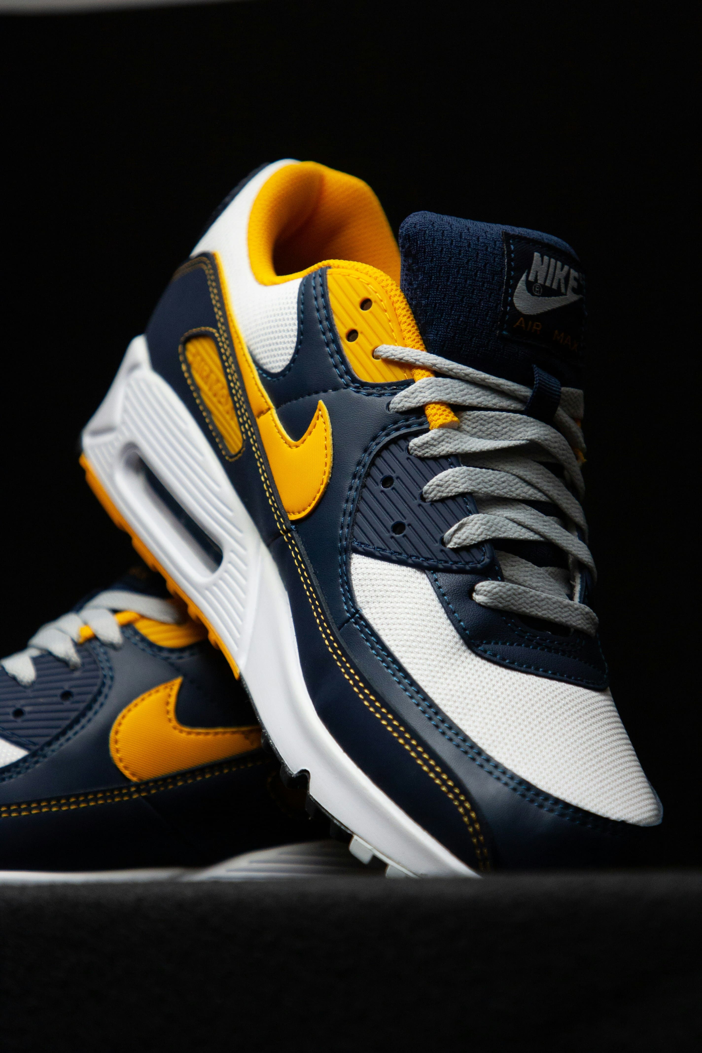 A close-up, studio shot of a pair of navy blue and yellow Nike Air Max sneakers against a black background. The sneakers feature white accents, grey laces, and detailed stitching.