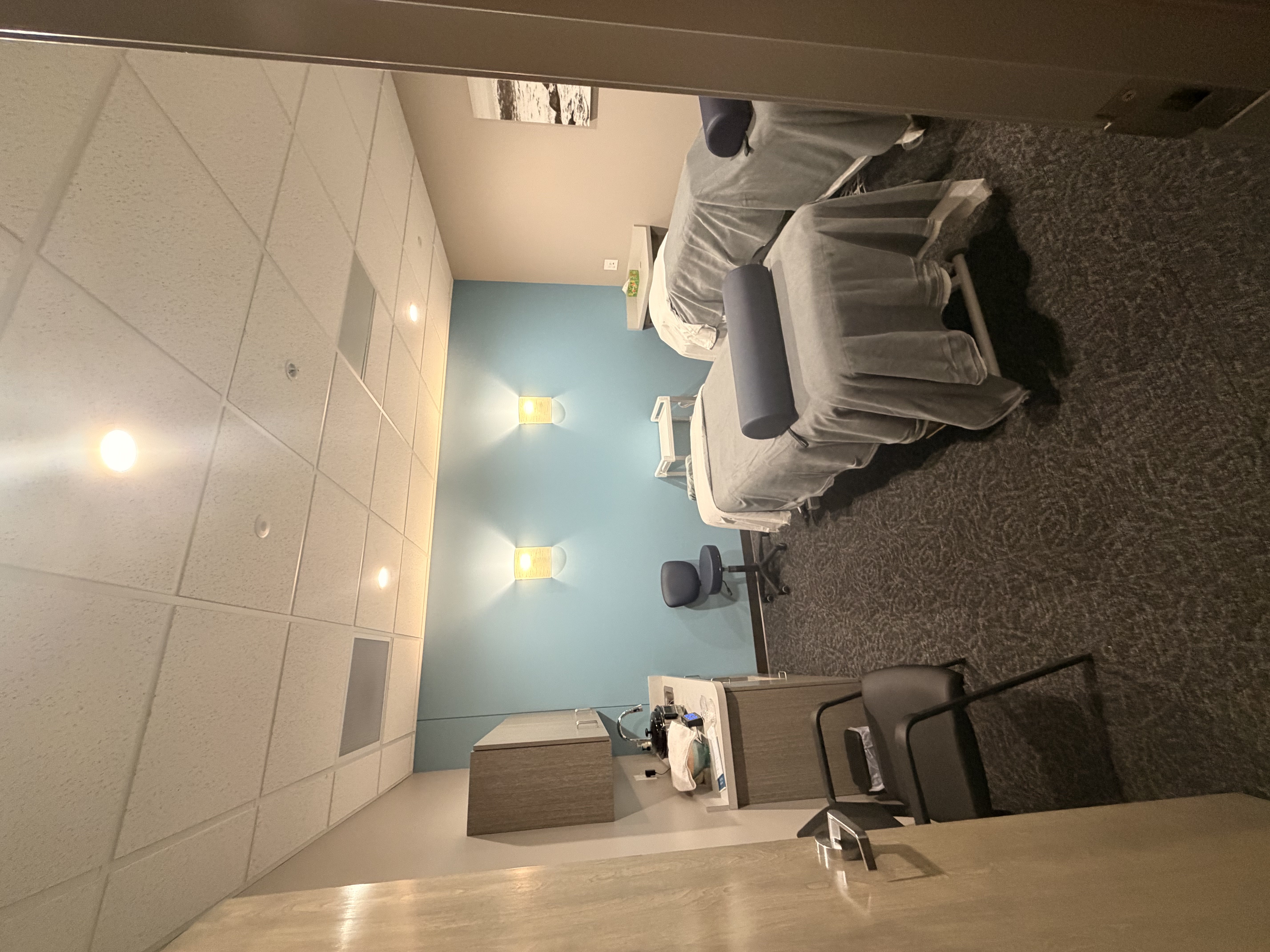 Dual massage treatment room with two massage tables covered in gray linens. Navy blue bolster pillows. Soft blue accent wall with wall-mounted sconce lighting. Gray carpet flooring and white ceiling with acoustic tiles. Treatment supplies on counter.
