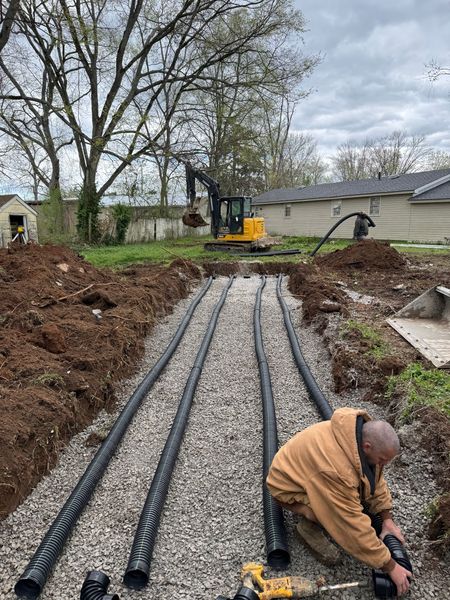 A worker in a brown jacket installs a septic drain field. Four long, black corrugated pipes are laid parallel in a gravel-filled trench, with an excavator operating nearby.