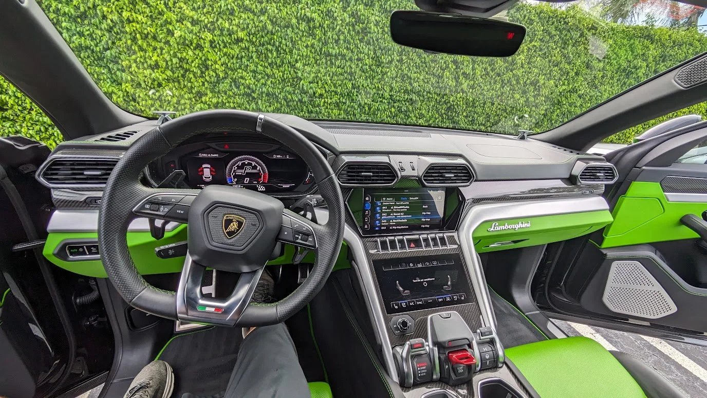 The interior of a luxury Lamborghini Urus SUV is showcased, featuring a striking design with neon green accents on the dashboard, doors, and seats. The steering wheel, adorned with the Lamborghini logo, and the central infotainment system with a touchscreen display are prominently visible. The car is parked outdoors, with lush green foliage serving as a backdrop.