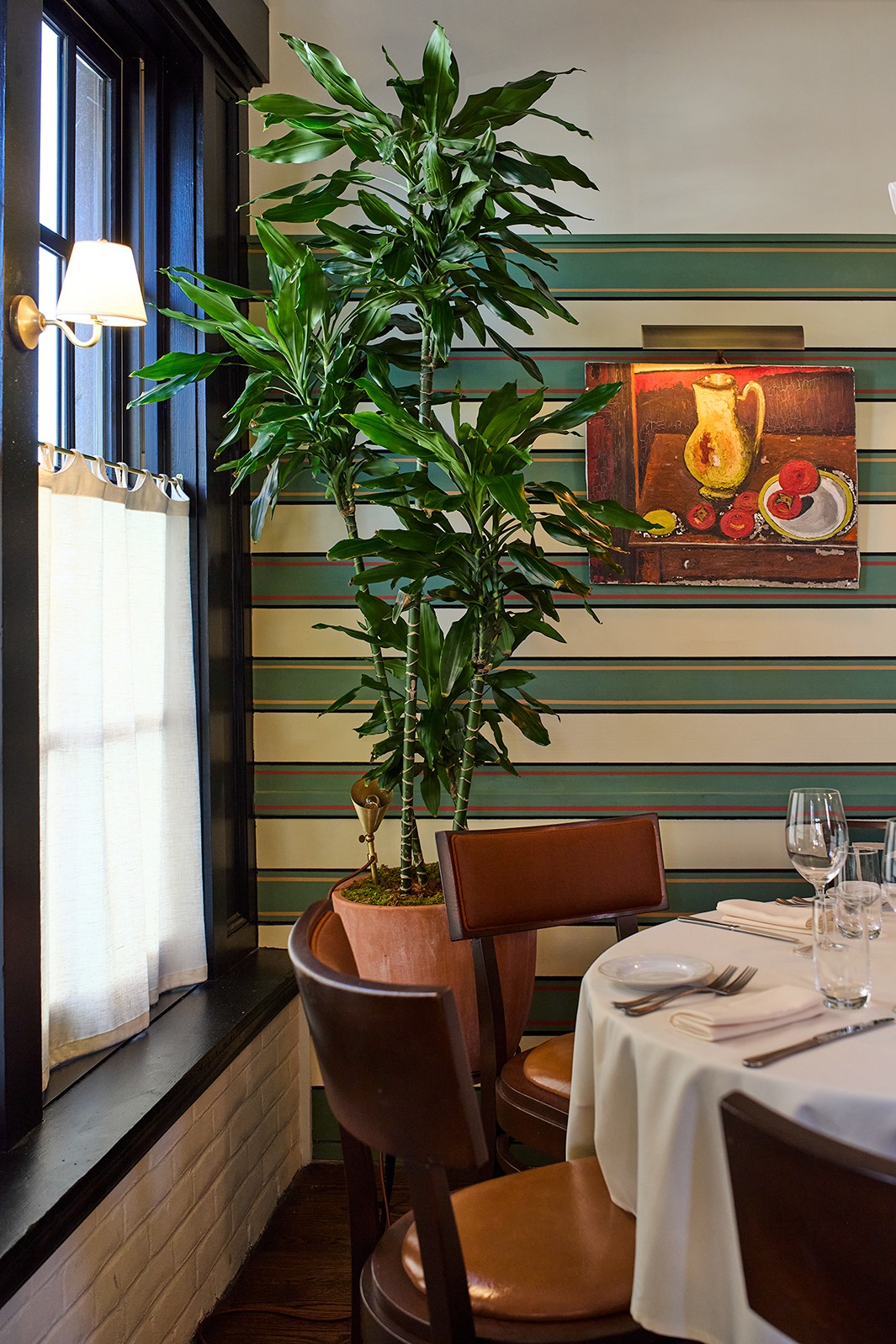 A dining table with white linens is tucked into a corner featuring green and tan striped wallpaper. A tall Dracaena plant in a terracotta pot stands next to a window and a framed still-life painting.
