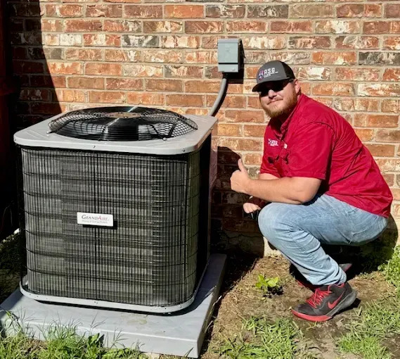 An HVAC technician in a red shirt and baseball cap crouches next to a new outdoor air conditioner condenser unit on a concrete pad, giving a thumbs up to the camera.