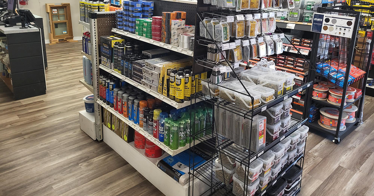 Interior showroom display featuring adhesives, caulks, and spray foam products on shelving alongside bins of fasteners and wire racks stocked with screws and nails, set on wood-look flooring near a checkout counter.