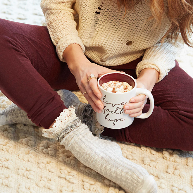 A woman sits on a plush white carpet in comfortable cable knit socks, burgundy leggings and a waffle knit beige sweater, she is holding a mug that says "Love, faith & hope" in cursive.