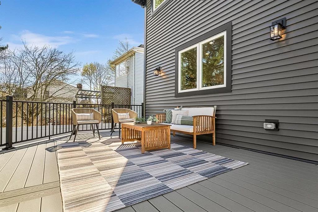 A modern outdoor deck with a wooden furniture set including a bench and two chairs, a wooden coffee table, and a striped rug. The deck is attached to a house with dark gray siding and a large window. There is a black metal railing around the deck, and beyond that, trees and neighboring houses are visible under a blue sky with scattered clouds.