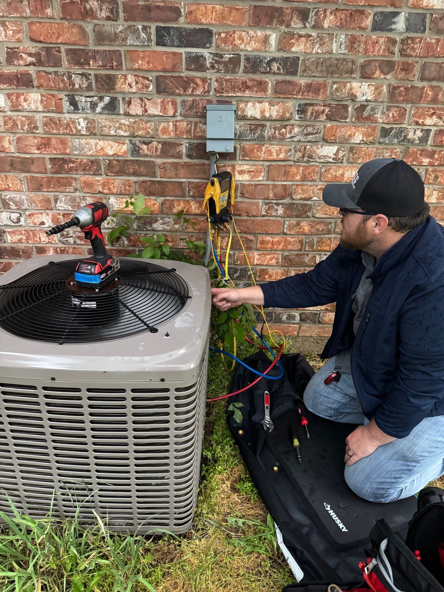 An HVAC technician wearing a baseball cap is kneeling beside an outdoor AC unit next to a brick wall, performing diagnostics with yellow and red gauges and tools.