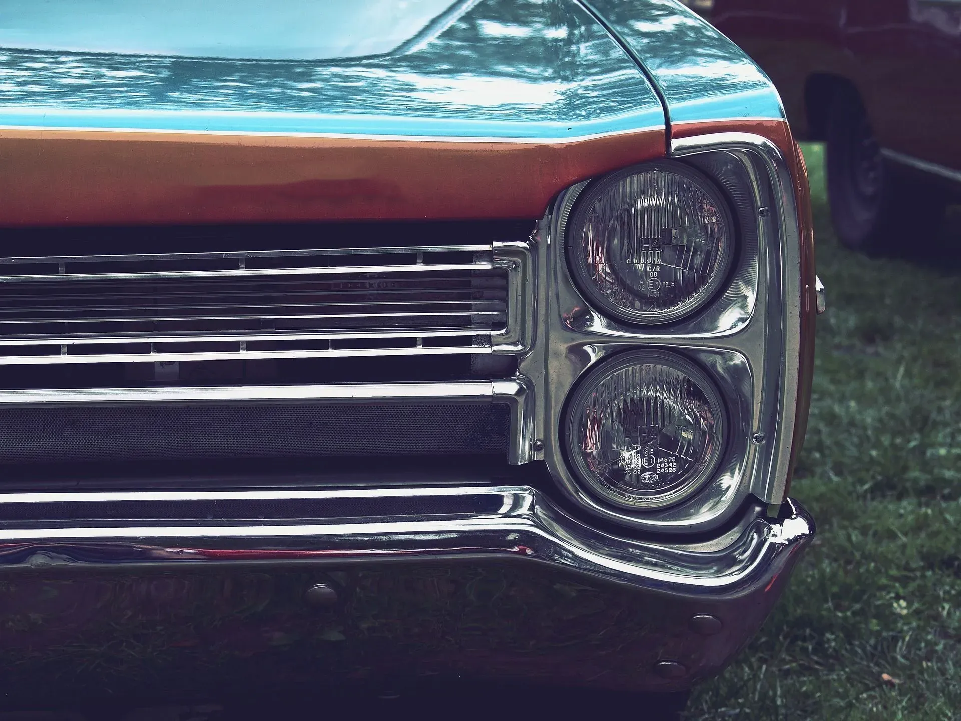 A close-up shot of the front of a classic car, featuring its dual headlights, chrome grille, and a two-tone paint job of teal and orange. The car is parked on a grassy lawn, and another car is visible in the background.