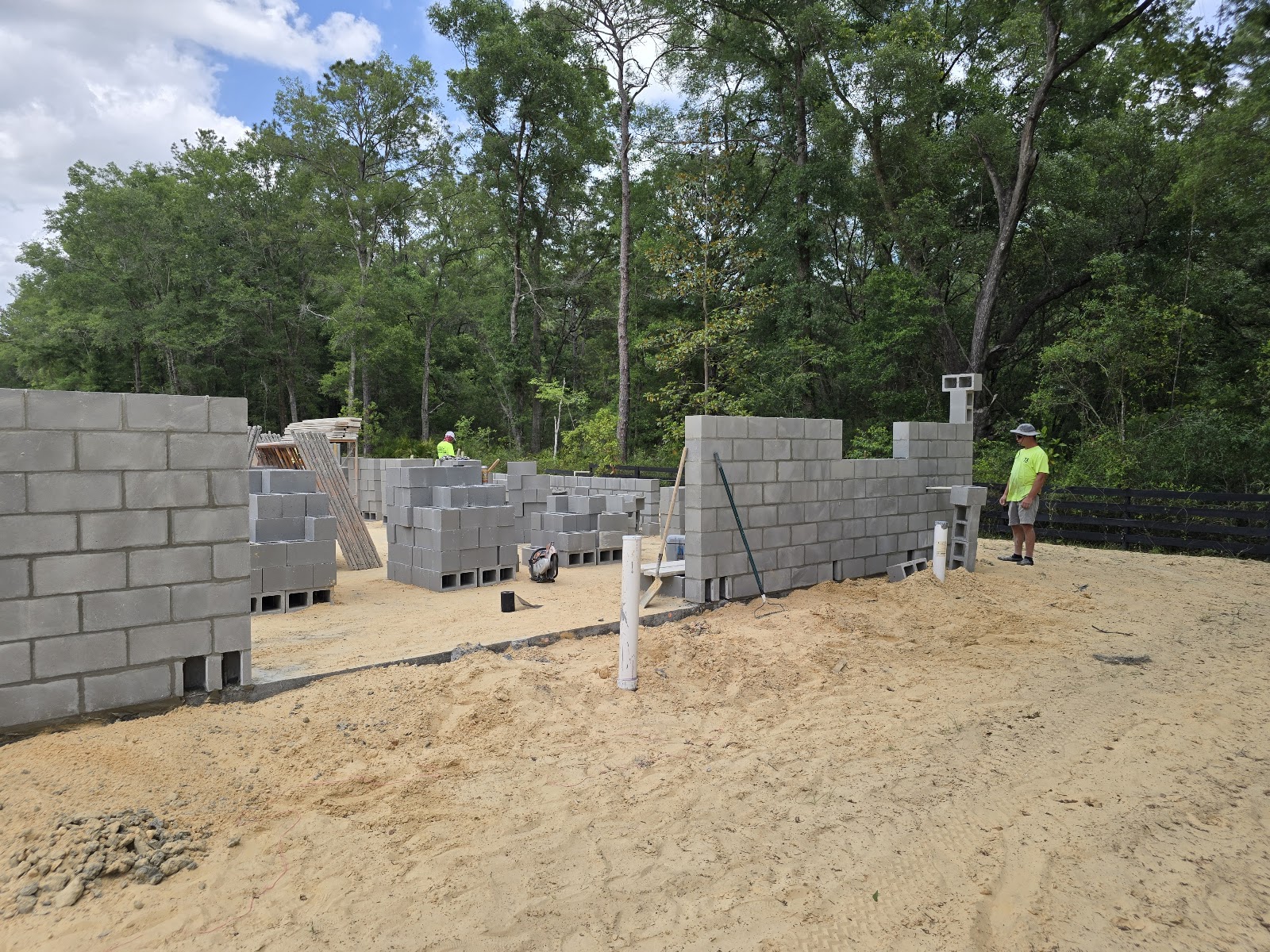 Construction workers are building a cinder block structure outdoors, surrounded by trees. The walls  