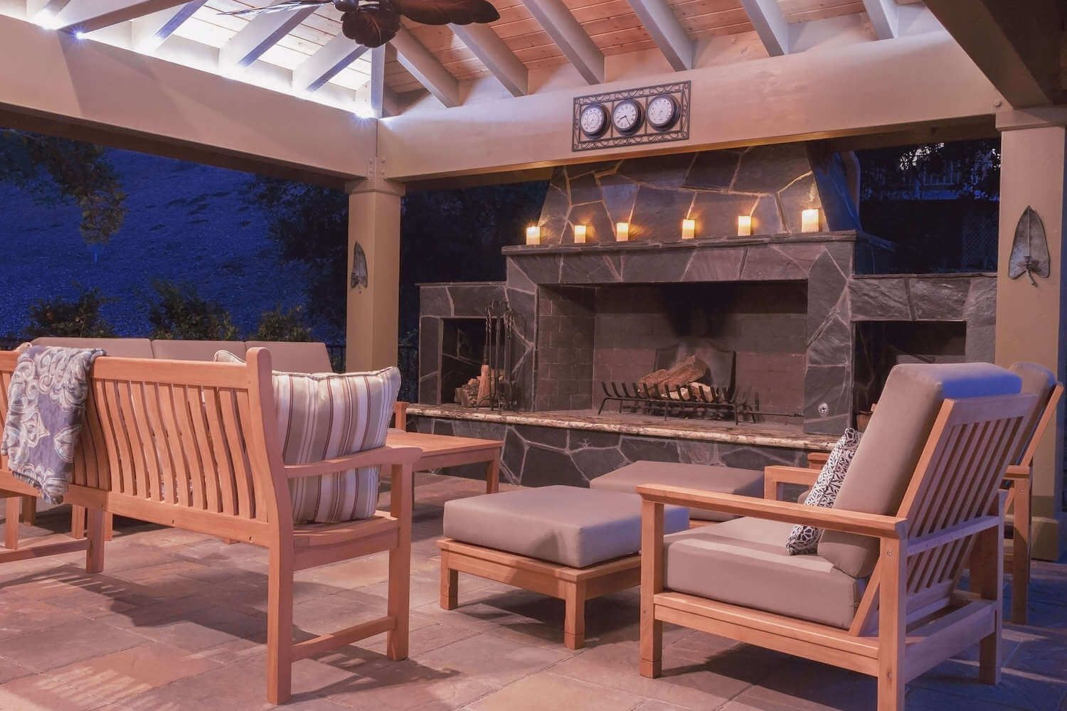 Cozy covered patio with a stone fireplace, glowing candles, cushioned wooden seating, and evening woods beyond; warm, rustic, inviting.