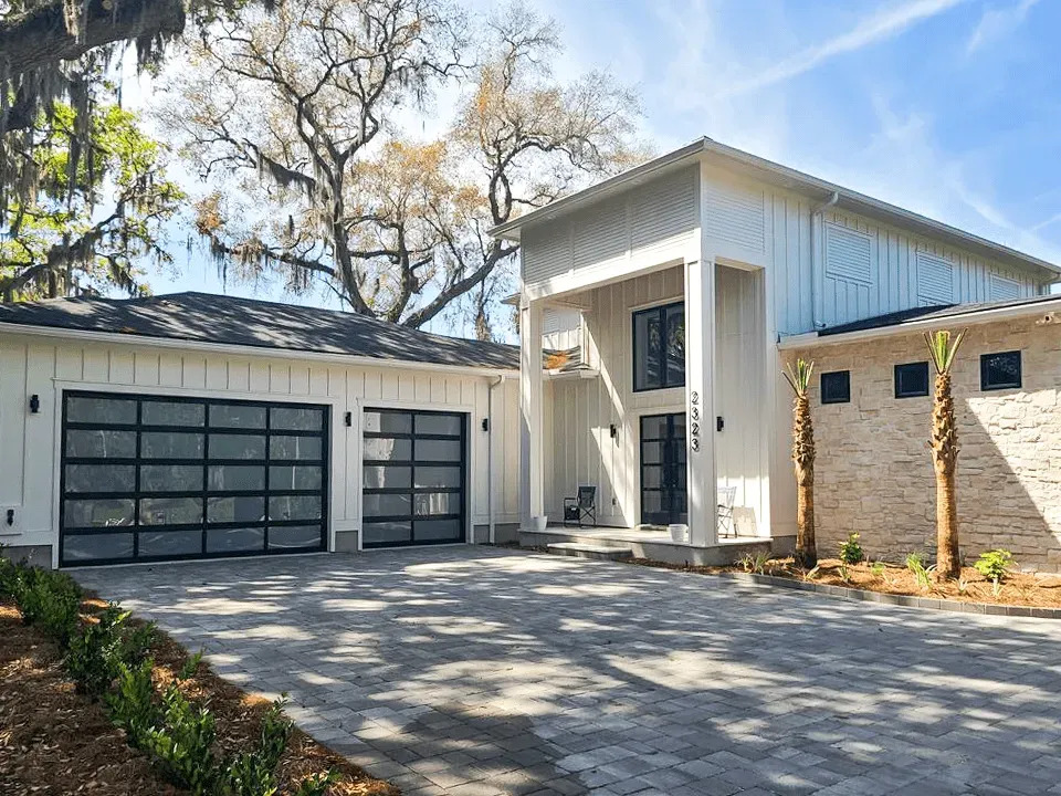 A modern residence showcases two black-framed glass garage doors with transparent panels, white siding, stone accents, landscaped beds, and a broad patterned paver driveway creating contemporary curb appeal and character.