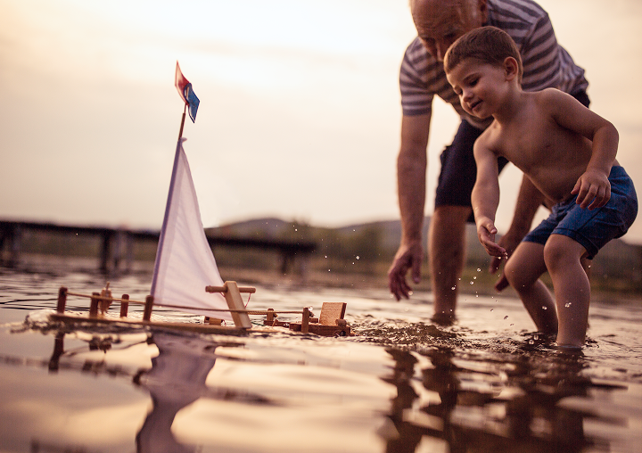 A grandfather and grandson playing with a model sailboat in a lake.