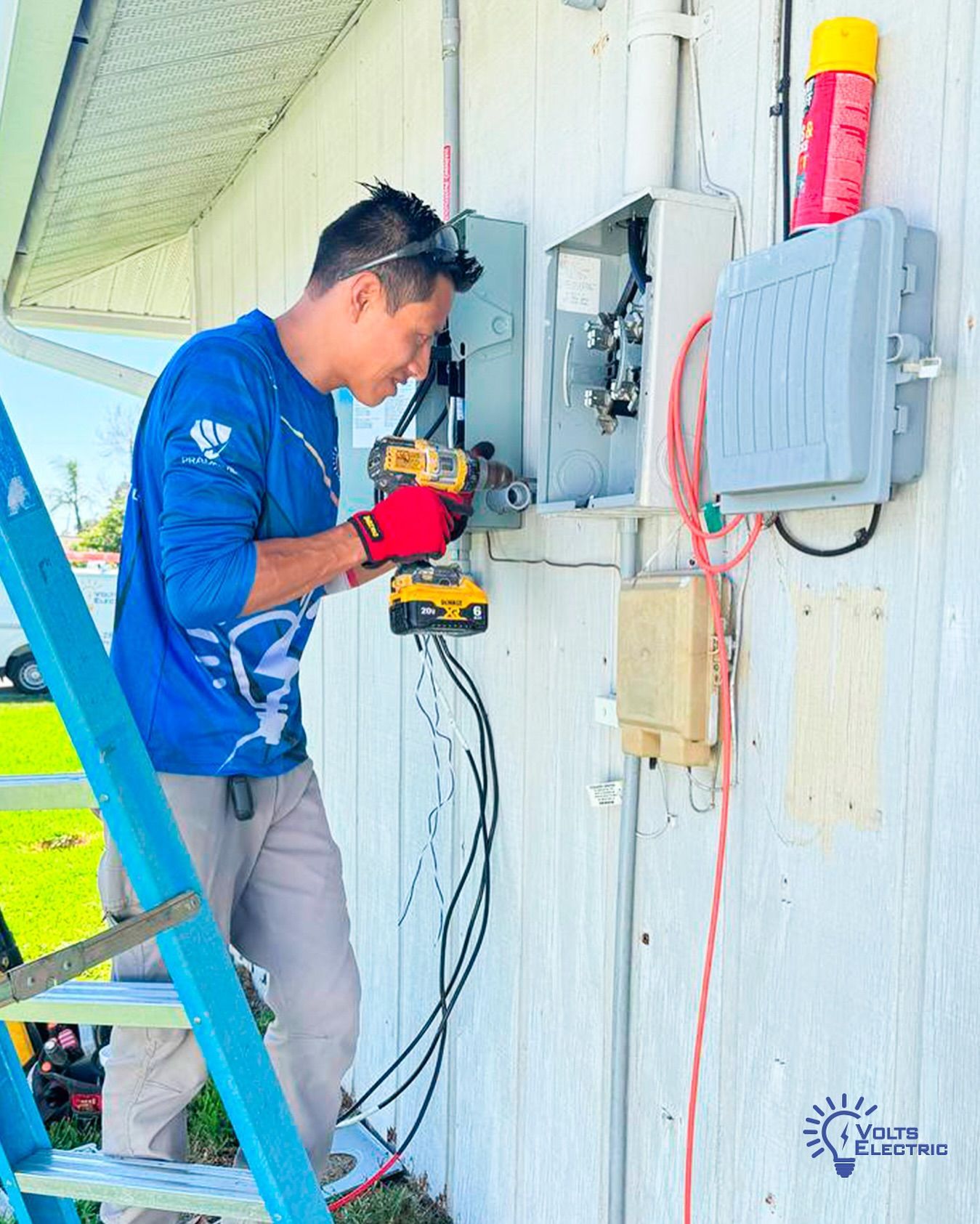 Electrician standing on a ladder installing or repairing an outdoor electrical service panel using a power drill, with exposed wiring and utility boxes mounted on the exterior wall.