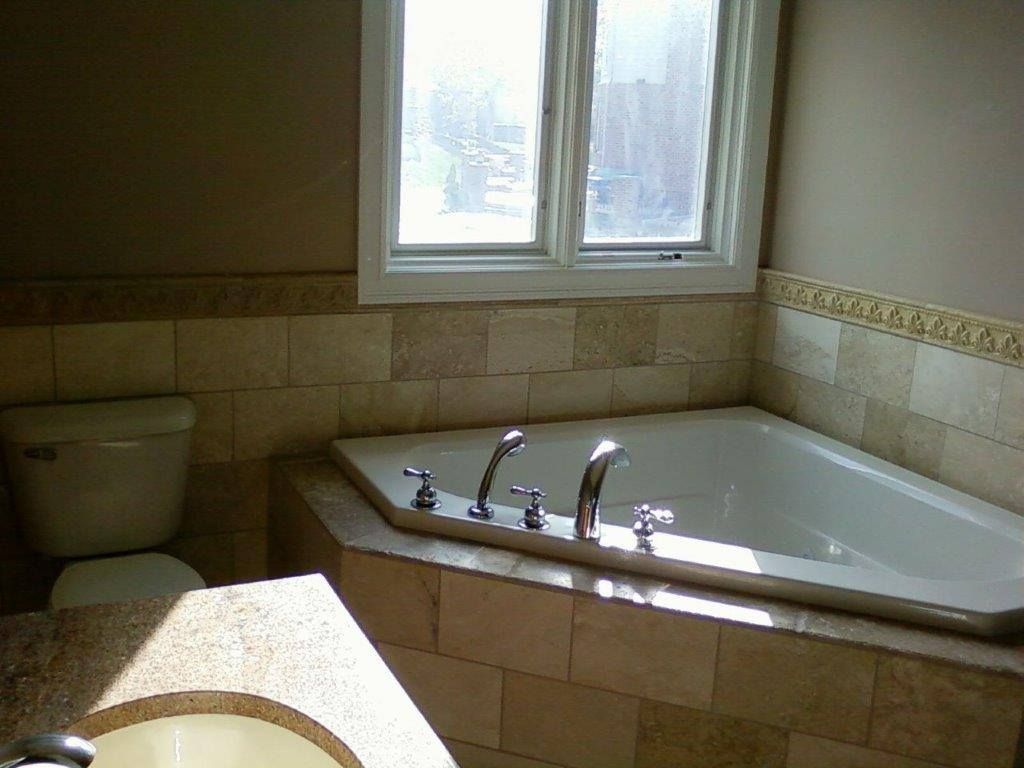 A bright bathroom featuring a built-in bathtub with tiled surround, chrome fixtures, and a nearby toilet. A large window provides natural light, enhancing the clean and functional design.