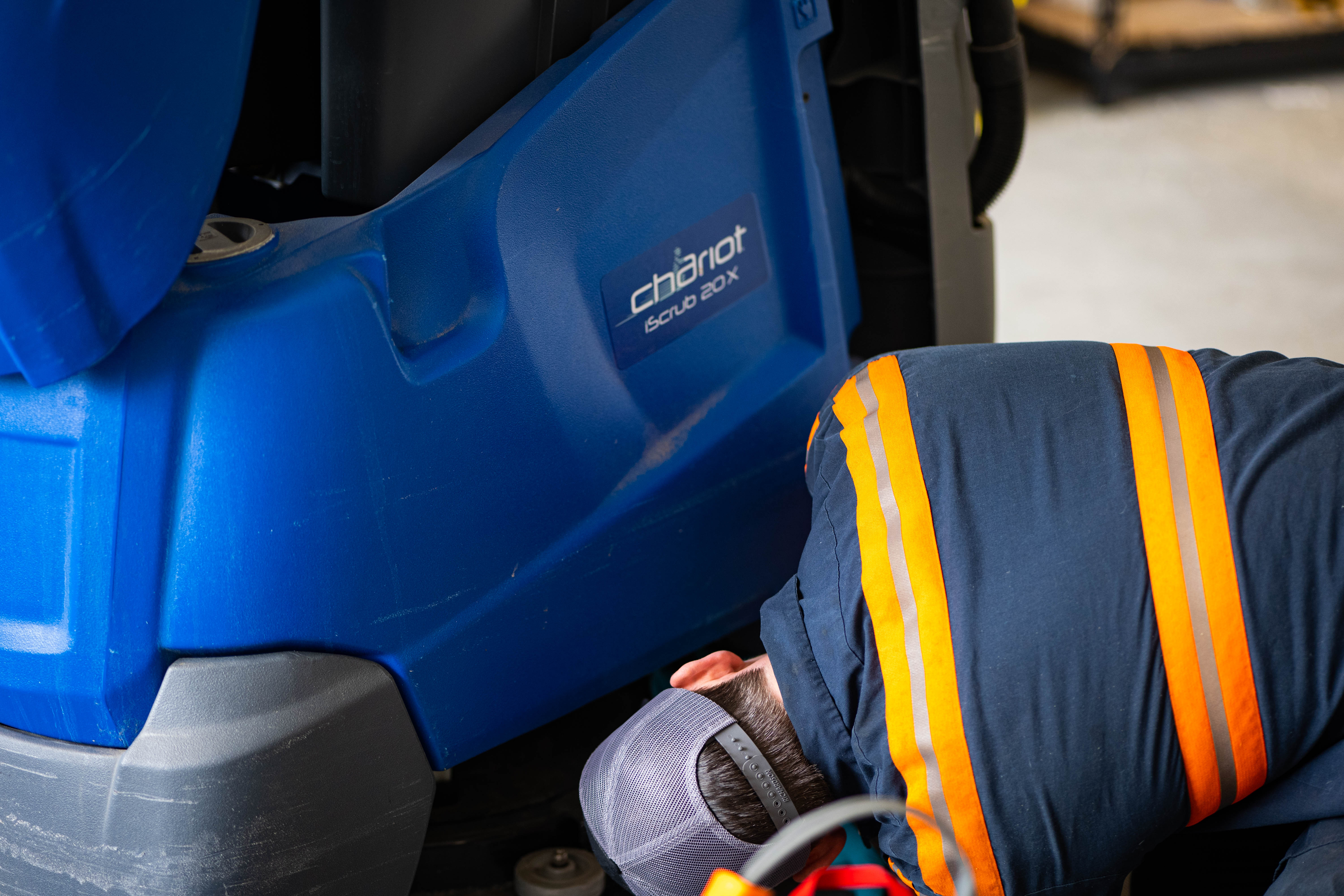 A WAY Solutions technician inspecting the lower brush deck, squeegee assembly, and drive motor of a blue Chariot iScrub 20X stand-on commercial floor scrubber.