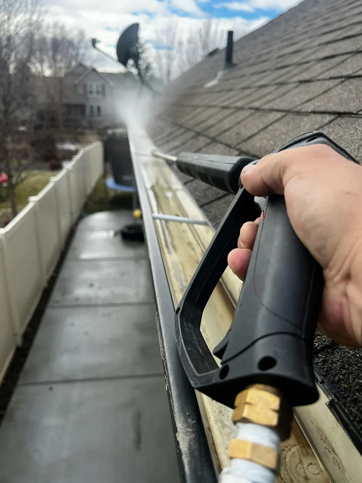 Close-up of a person using a pressure washer to clean roof shingles and gutters, removing dirt, debris, and buildup. High-pressure water spray improves cleanliness, prevents damage, and helps maintain the home&rsquo;s exterior condition and overall appearance.