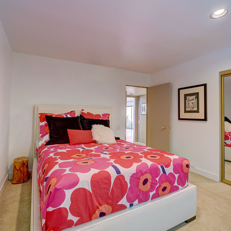 A bedroom with a large bed featuring a vibrant floral duvet cover in shades of pink and red. Several throw pillows, including black velvet and a fluffy white one, are arranged on the bed. To the left of the bed is a small wooden tree stump serving as a side table. The room has white walls, a light-colored carpet, and a framed picture on the wall opposite the bed. A doorway leads to another part of the house.