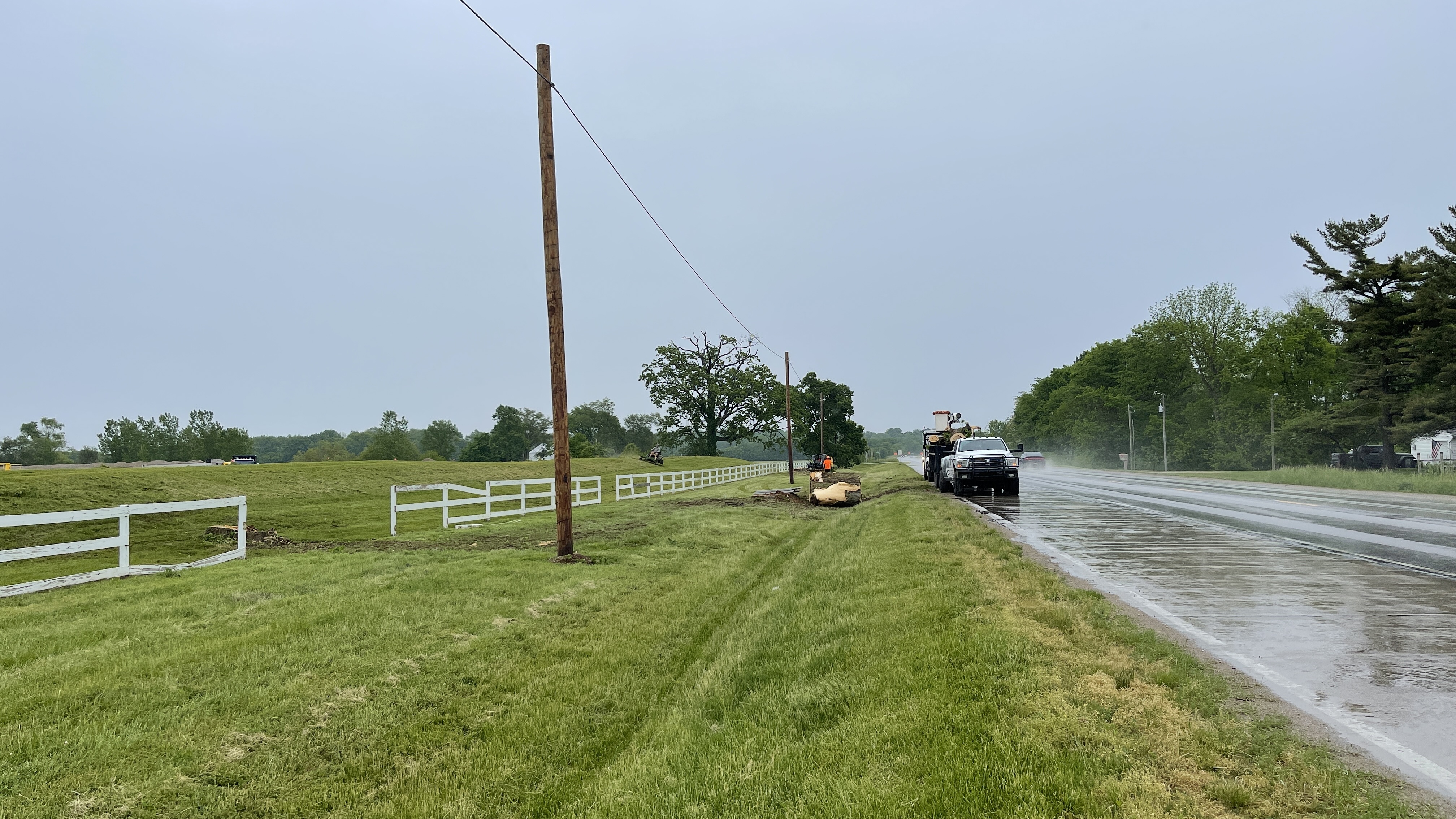 A utility truck is parked on the side of a wet road next to a field with a white fence. A worker in an orange vest is standing near a large log. Power lines are visible in the background, strung between wooden poles. The sky is overcast and it appears to be raining.