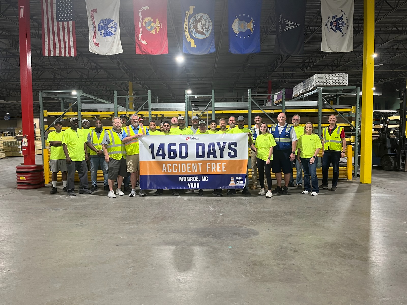 Group of Builders FirstSource team members standing inside a warehouse holding a banner that reads “1460 Days Accident Free, Monroe, NC Yard.” Team members wear high-visibility safety apparel, with warehouse racking, building materials, forklifts, and U.S. military branch flags visible in the background.