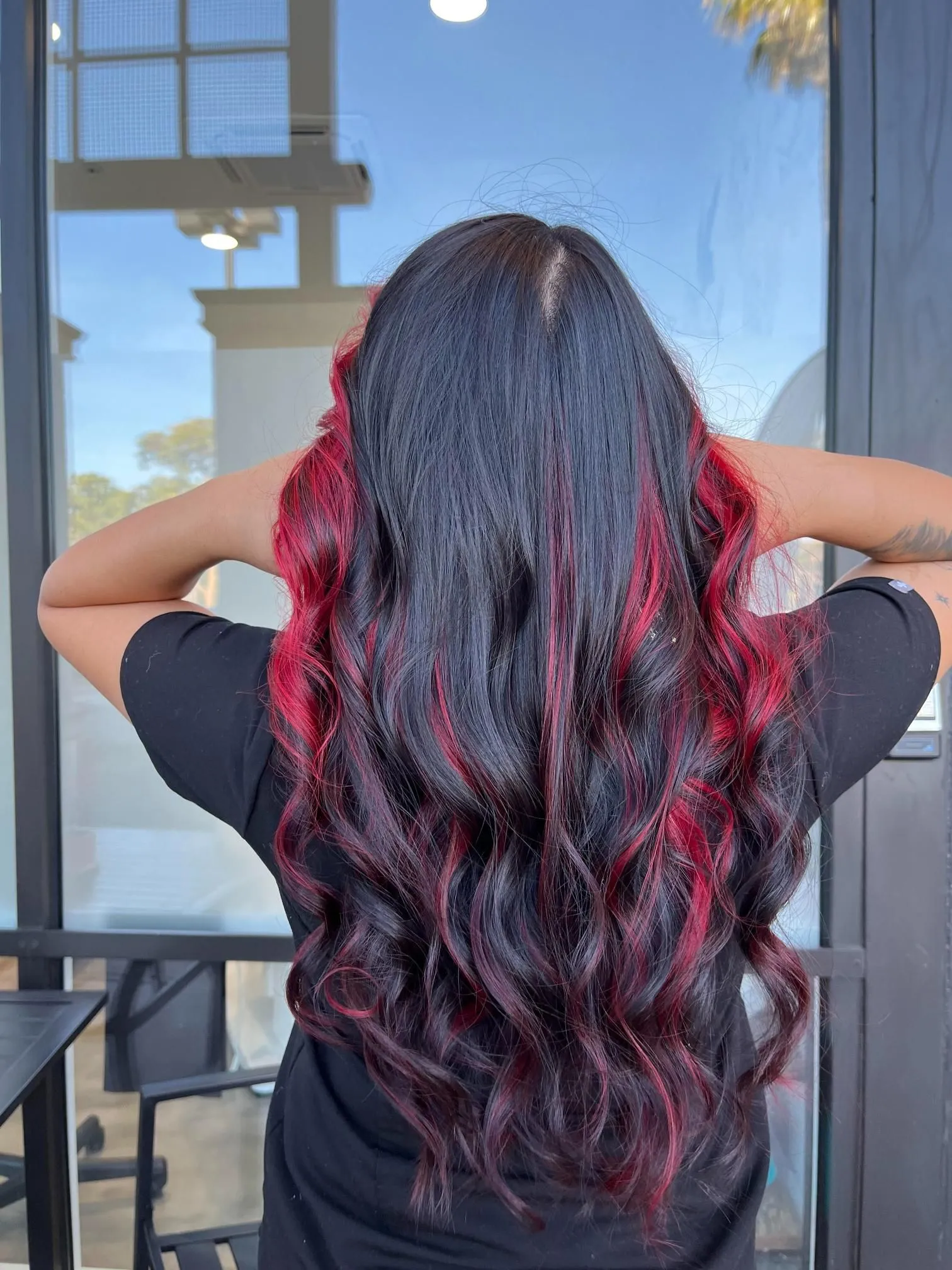 Long black hair with bold red highlights styled in soft flowing curls, creating strong contrast and dimension. The hairstyle showcases creative salon coloring techniques and professional styling from the back.