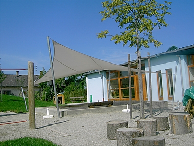 graues, gespanntes Sonnensegel "Drachenstein", über dem Außenbereich eines kleinen, blauen Kindergarten.