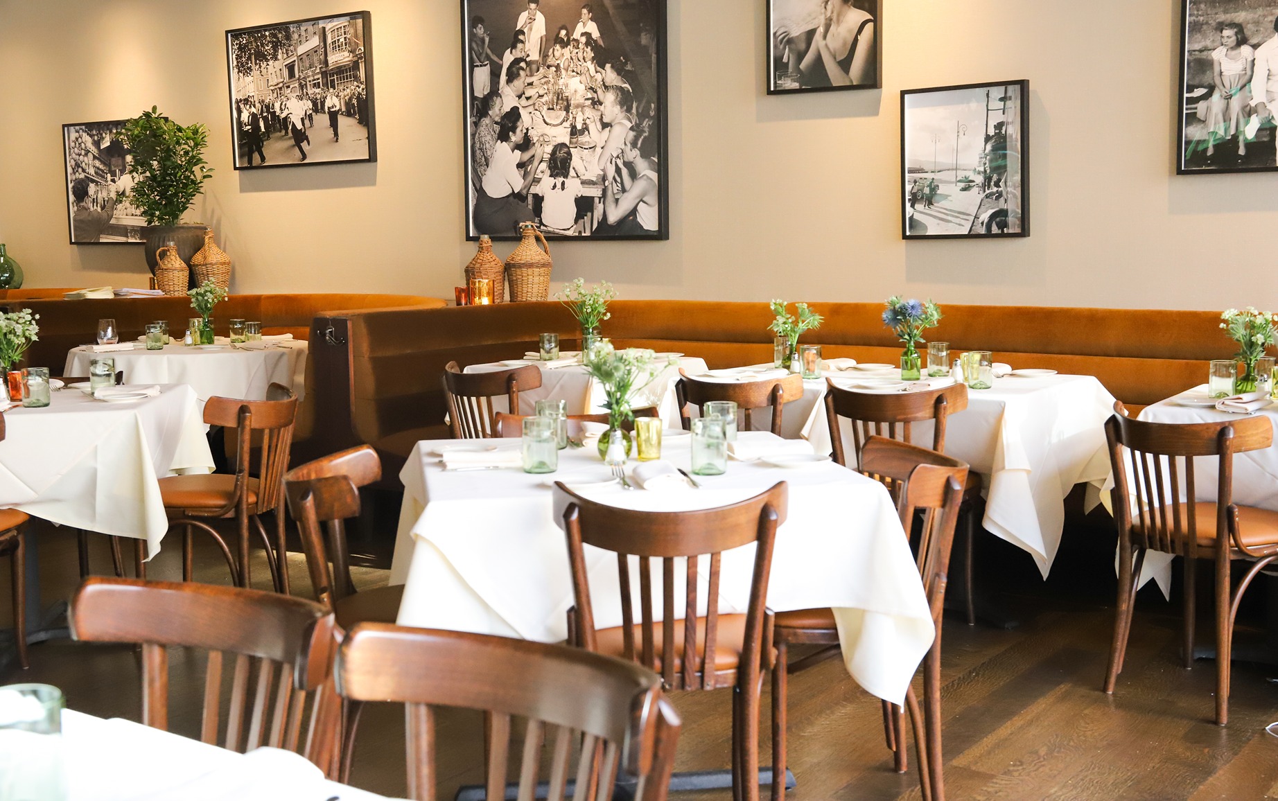 The elegant main dining area featuring white linen tables, classic wooden chairs, and a long tan leather banquette. Walls are decorated with large vintage Italian photos.
