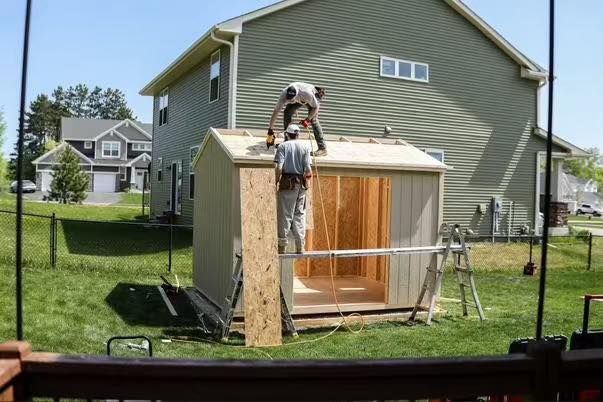 Two construction workers are building a small, unpainted shed with a gable roof in a suburban backyard. One worker is securing the roof decking while the other stands inside the structure, with a green-sided house in the background.
