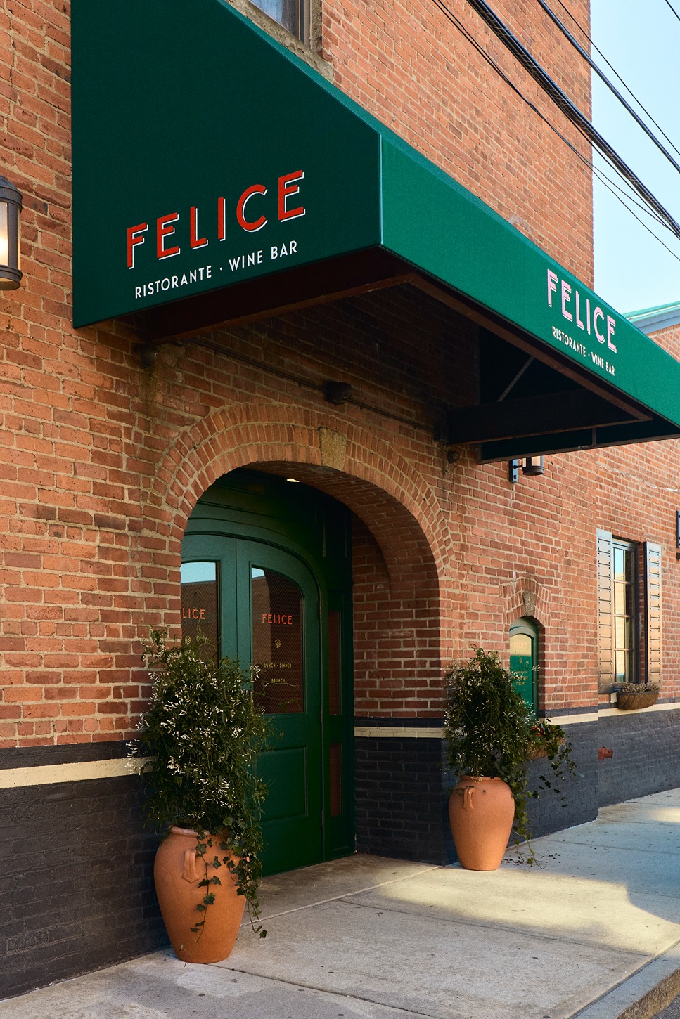 The storefront of Felice featuring dark wood framing, large glass windows, and signature forest green awnings. Outdoor bistro seating is visible on the sidewalk under a clear blue sky.