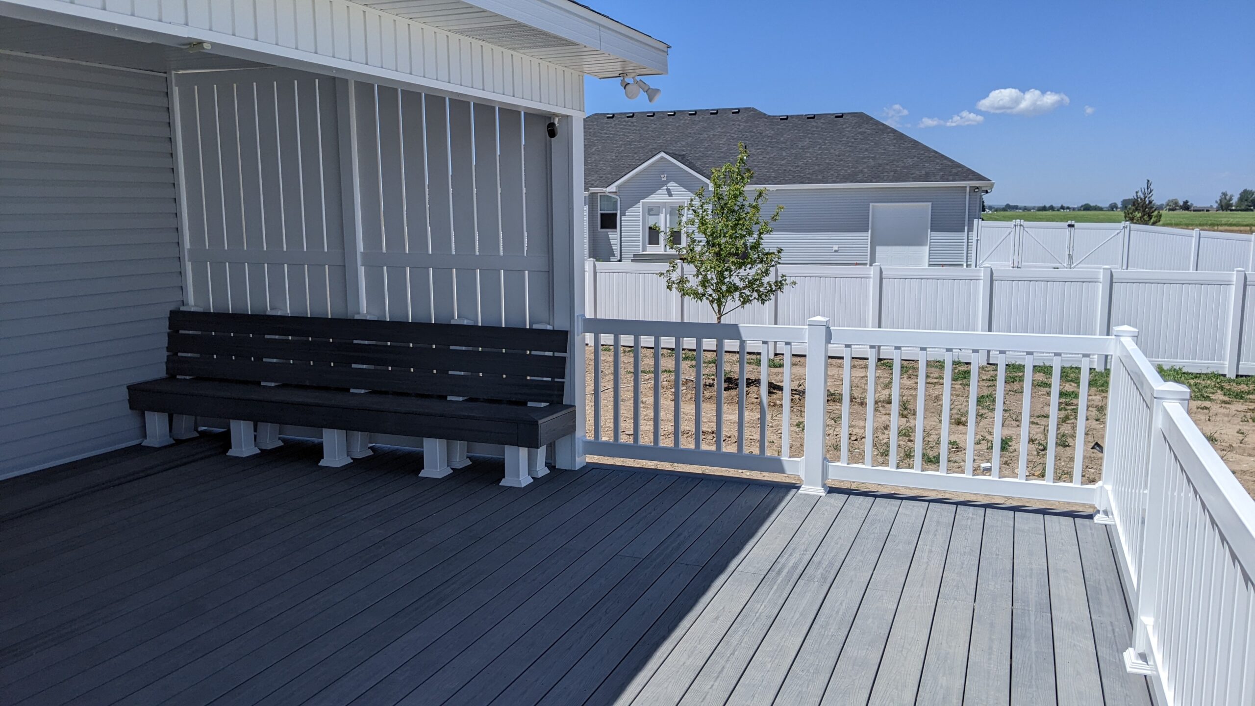 Expand your outdoor living possibilities with this extensive multi-level deck project by DeckPro LLC. Pictured on a sunny day, this build features high-quality composite decking in a warm wood-grain finish and contrasting dark perimeter trim. For the ultimate in style and function, we installed highly detailed black metal railings with unique curved balusters and wide, inviting stairs. The raised structure integrates seamlessly with natural rock retaining wall landscaping, leading down to a lower-level concrete patio for a truly multi-functional backyard retreat.