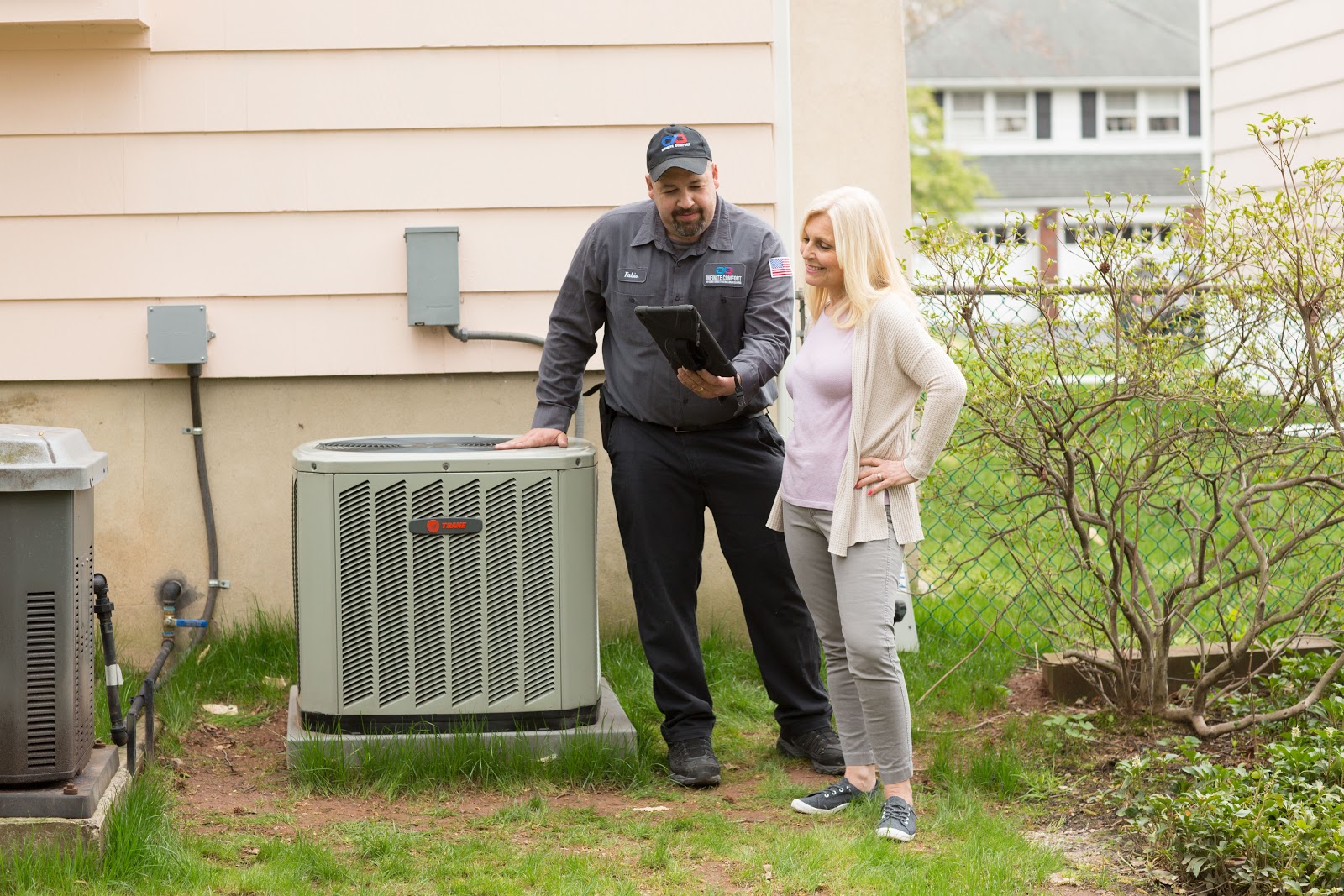 A HVAC technician stands next to an outdoor air conditioning unit, holding a tablet and discussing something with a homeowner. The technician is wearing a gray uniform with a logo, and the homeowner is wearing a light purple shirt and a beige cardigan. They are standing in a residential yard with a light-colored house in the background.