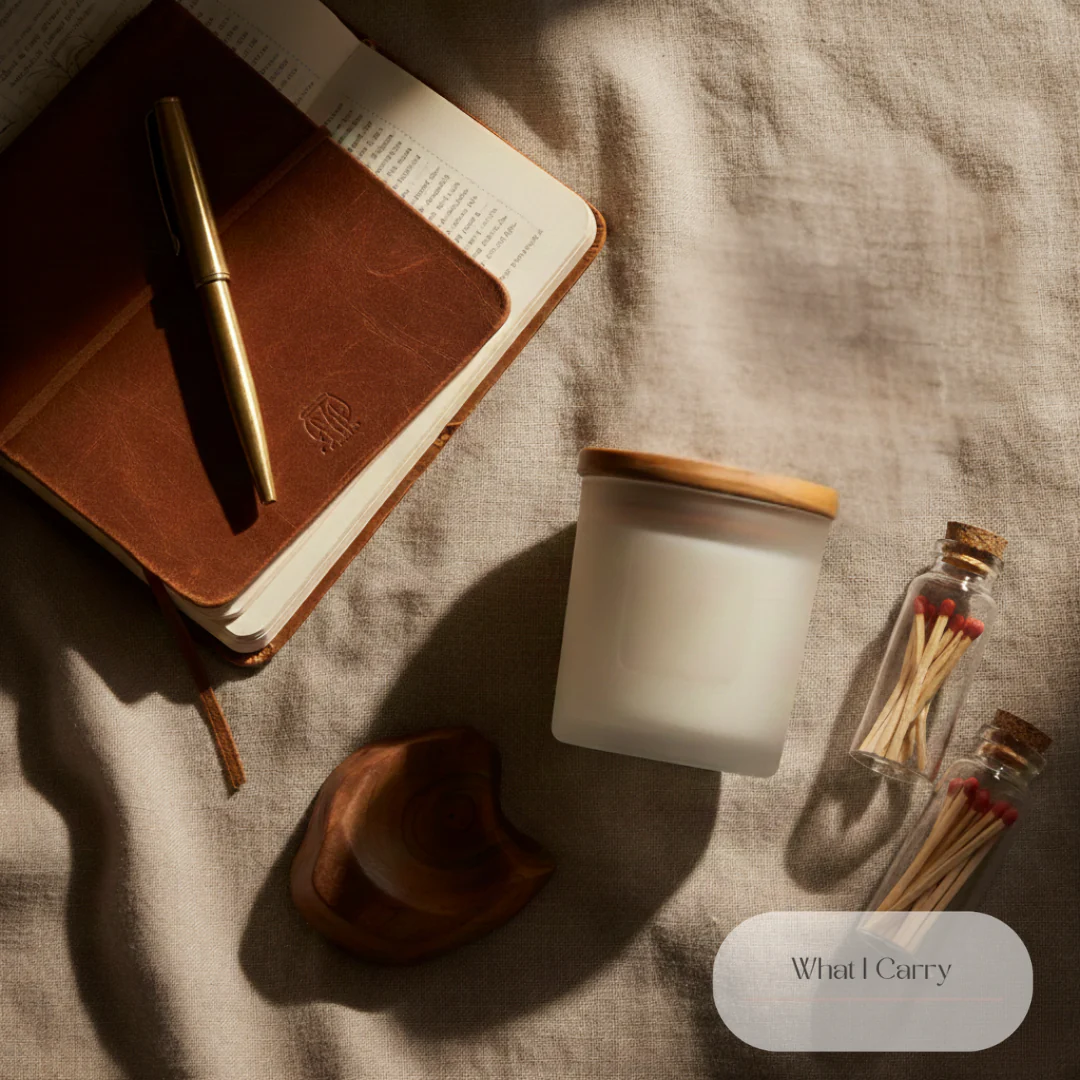 A flat lay composition of lifestyle items includes a leather-bound notebook with a gold pen resting on it, a frosted glass candle with a wooden lid, a small carved wooden object, and two small glass vials filled with matches. The items are arranged on a textured linen fabric, casting soft shadows. A subtle oval banner at the bottom right displays the text "What I Carry."