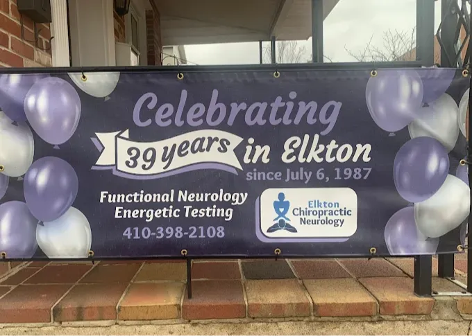 It&rsquo;s a photo of a large outdoor banner hung along a porch/railing, with a dark purple background and balloon graphics around the edges (purple, white, and silver).
The banner text reads &ldquo;Celebrating 39 years in Elkton&rdquo; and includes &ldquo;since July 6, 1987.&rdquo; Below that it lists services: &ldquo;Functional Neurology&rdquo; and &ldquo;Energetic Testing,&rdquo; plus the phone number 410-398-2108. On the right side there&rsquo;s a logo box that says &ldquo;Elkton Chiropractic Neurology&rdquo; with a small meditating figure icon.