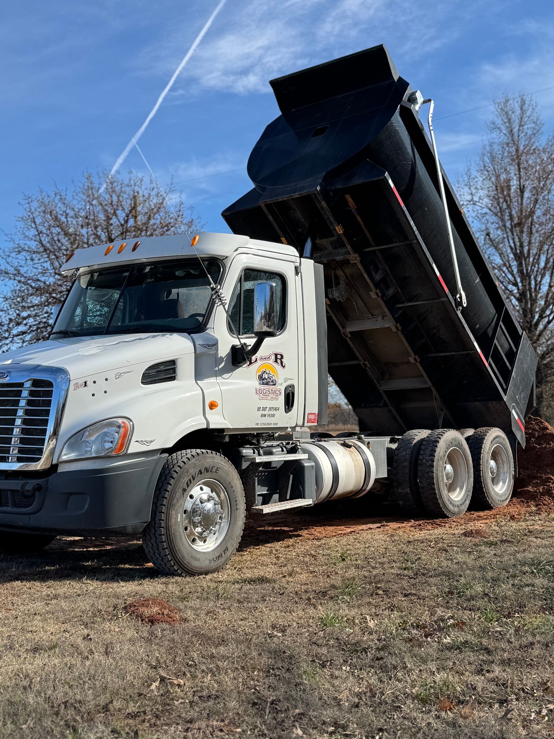 L & R Logistics LLC dump truck with raised black bed parked outdoors under a bright blue sky, showcasing heavy-duty hauling equipment and professional transportation capabilities for local Oklahoma projects.