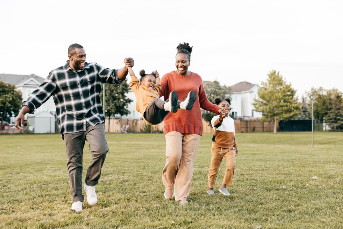 A Black family of four, two parents and two young daughters, are walking and playing on a grassy field with houses and trees in the background under an overcast sky. The father, on the left, is wearing a black and white plaid shirt and grey pants, holding his daughter's hand while she swings in the air. The mother, in the center, is wearing a rust-colored sweater and tan pants, holding her other daughter's hand. The daughter on the right is wearing a brown and white color-blocked sweater and tan pants.