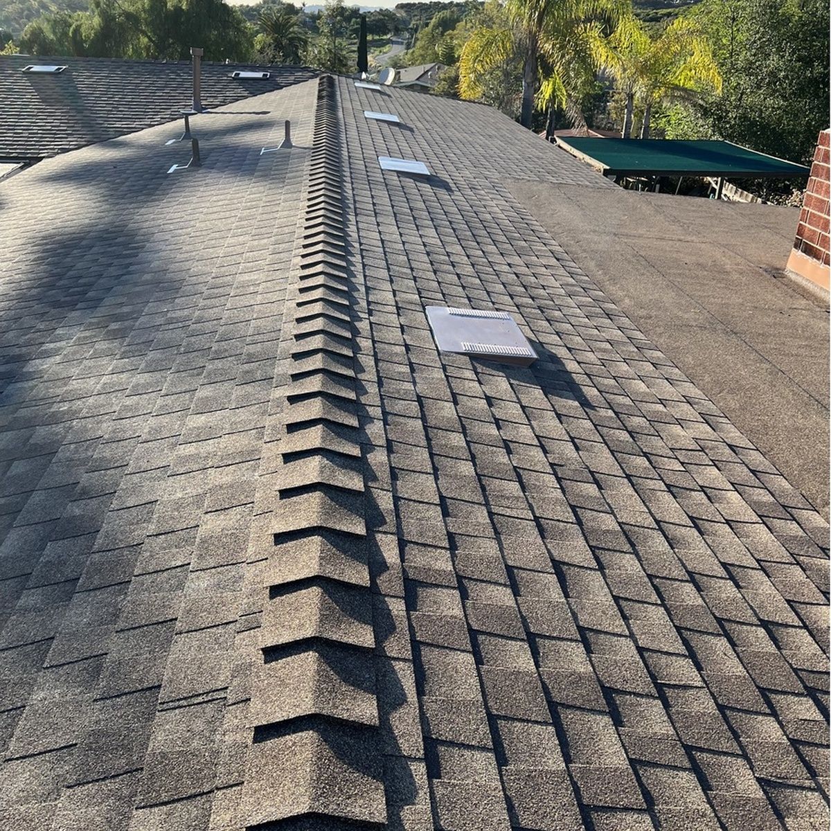 Perspective shot following the central ridge line of a dark gray asphalt shingle roof, featuring rectangular attic vents, roof pipes, and neighboring greenery in the distance.