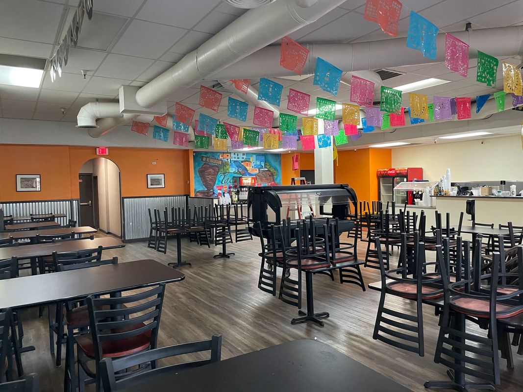 A colorful restaurant interior with tables and chairs stacked for cleaning, decorated with vibrant hanging banners, featuring a service counter and beverage area, creating a lively and casual dining atmosphere.