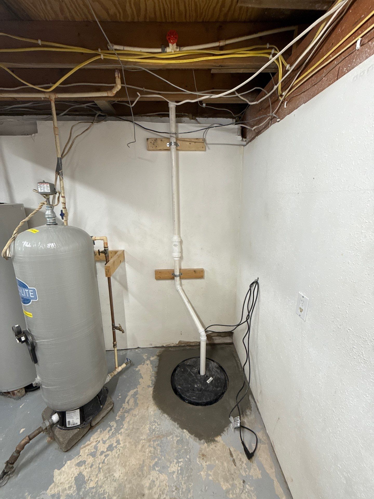 Basement sump pump installation includes a covered floor basin, vertical PVC discharge pipe, pressure tank, exposed ceiling wiring and plumbing, white walls, and worn painted concrete flooring around the equipment.