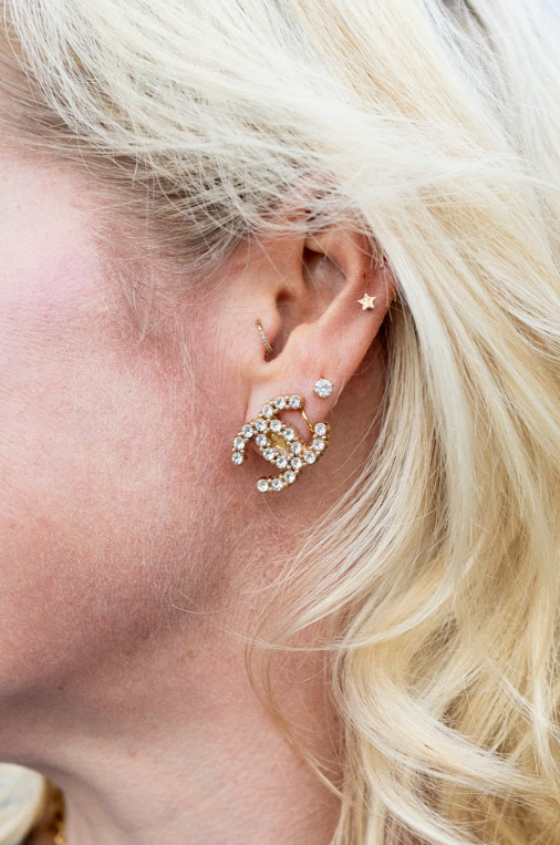 A sparkling gold-toned logo earring pairs with a clear stone stud and little star stud for a layered look. Discover statement earrings and curated jewelry at Eliana Curated in Old Town Alexandria, VA.