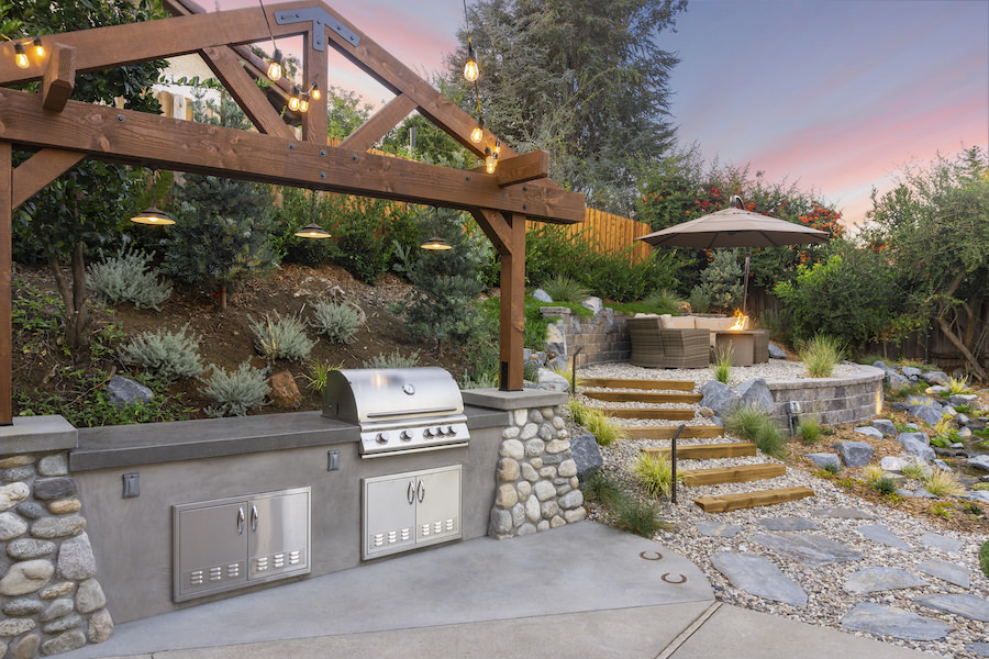 Outdoor backyard kitchen with stainless-steel grill, pergola string lights, stone patio, garden steps, umbrella seating, and a pink sunset.
