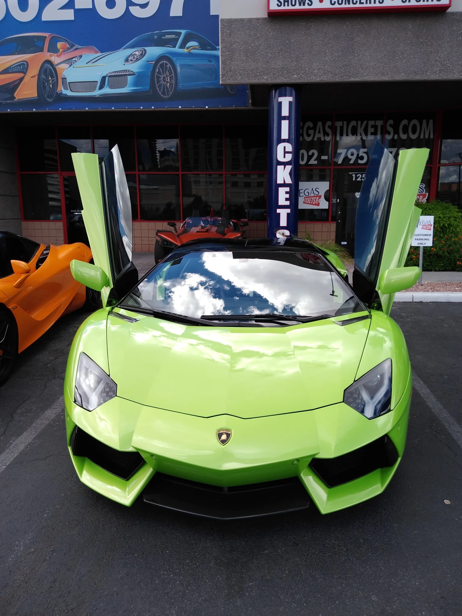 A bright lime green Lamborghini with its scissor doors open is parked in a lot in front of a building that advertises ticket sales for shows, concerts, and sports. The car's glossy finish reflects a cloudy sky. To the left, an orange sports car is visible, and in the background, a red vehicle with a similar aggressive design is parked. The building features large windows and a sign that reads "VEGASTICKETS.COM".