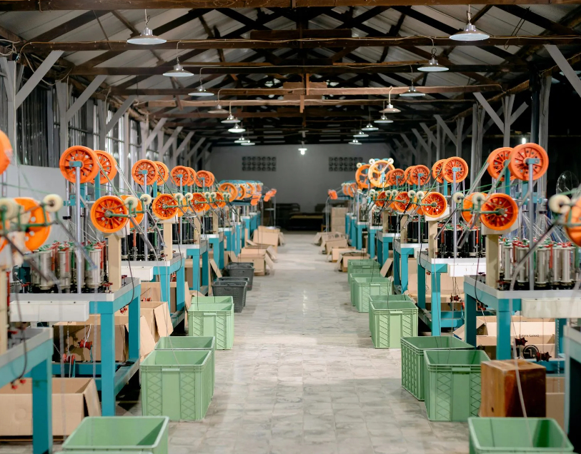 A high-angle interior view down the main aisle of a textile manufacturing or assembly plant, lined with rows of machinery, orange spool wheels, hanging pendant lights, and green storage bins.